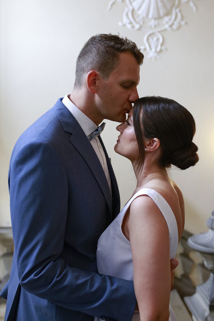 Braut und Bräutigam stehen sich nahe, umgeben von eleganter Architektur. Bräutigam küsst die Braut liebevoll auf die Stirn.