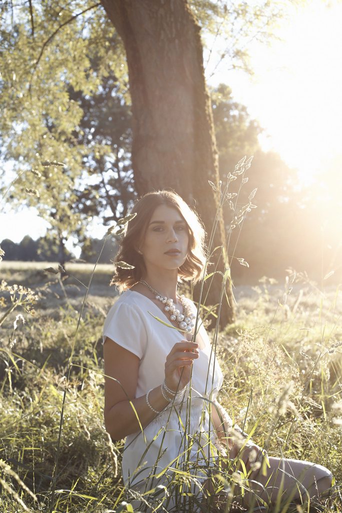 Frau in weißem Kleid sitzt in einer Wiese im Sonnenlicht. Hinter ihr befindet sich ein Baum.
