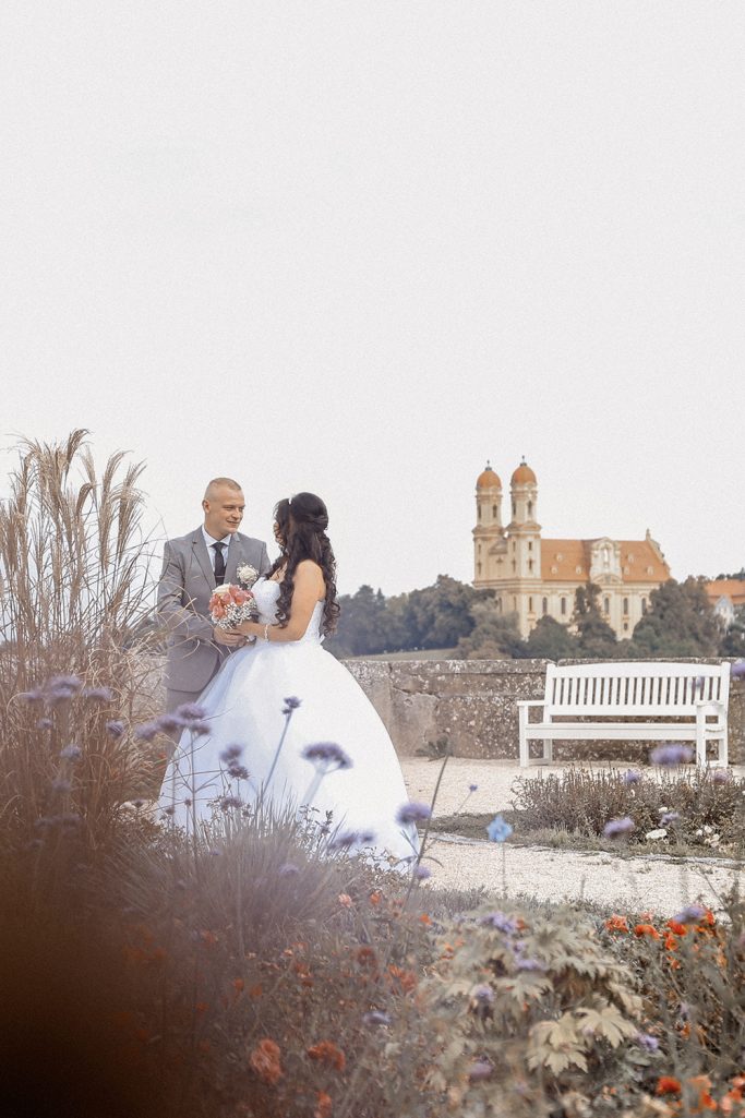 Brautpaar in einem Garten mit einer Kirche im Hintergrund, umgeben von Blumen.