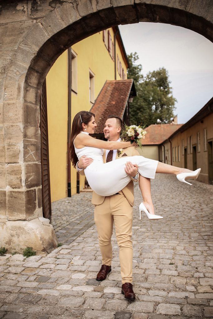 Brautpaar in einer Altstadt auf einem Kopfsteinpflasterweg. Er trägt die Braut auf den Armen.