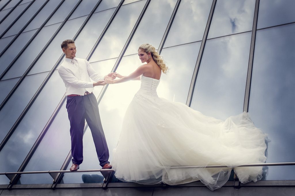 Braut und Bräutigam stehen auf einem Dach, umgeben von Wolken und Spiegelungen des Glasdachs.