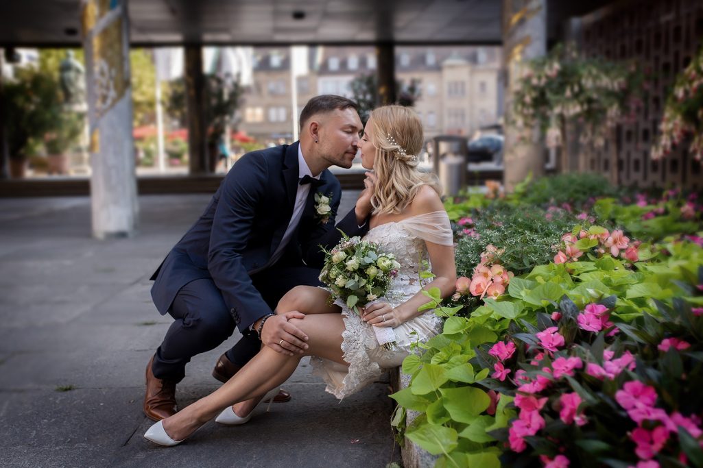 Brautpaar küsst sich vor blühenden Pflanzen, umgeben von urbaner Kulisse.