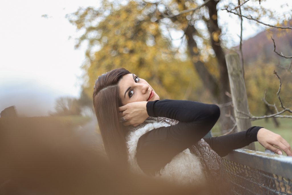 Junge Frau mit langen Haaren, die lächelnd vor herbstlichen Bäumen posiert.