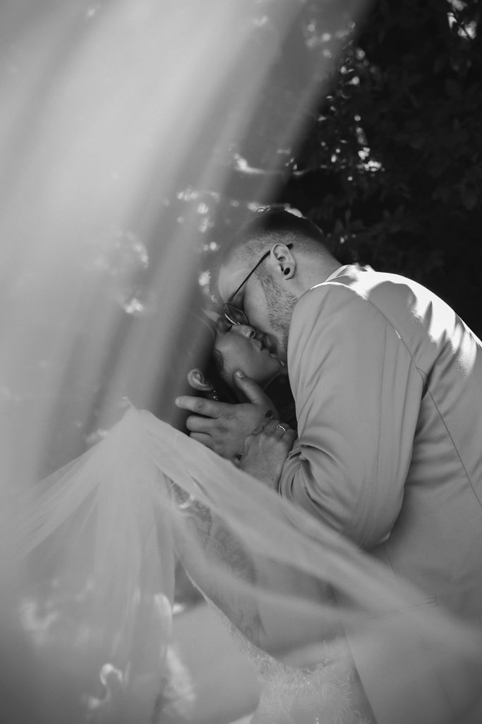 Ein Brautpaar küsst sich unter einem zarten, weißen Schleier, umgeben von Bäumen.