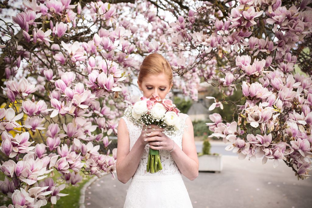Braut mit Blumenstrauß steht vor blühenden Magnolienbäumen.