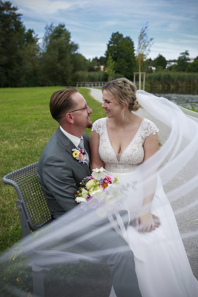 Ein Brautpaar sitzt lächelnd und sich gegenseitig anschauend auf einer Bank, umgeben von Grün und mit Schleier im Wind.