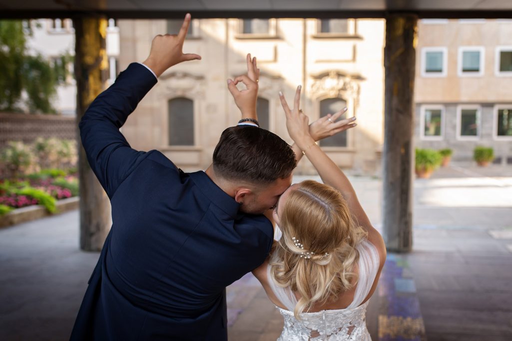 Braut und Bräutigam feiern mit Armbewegungen vor historischem Hintergrund.