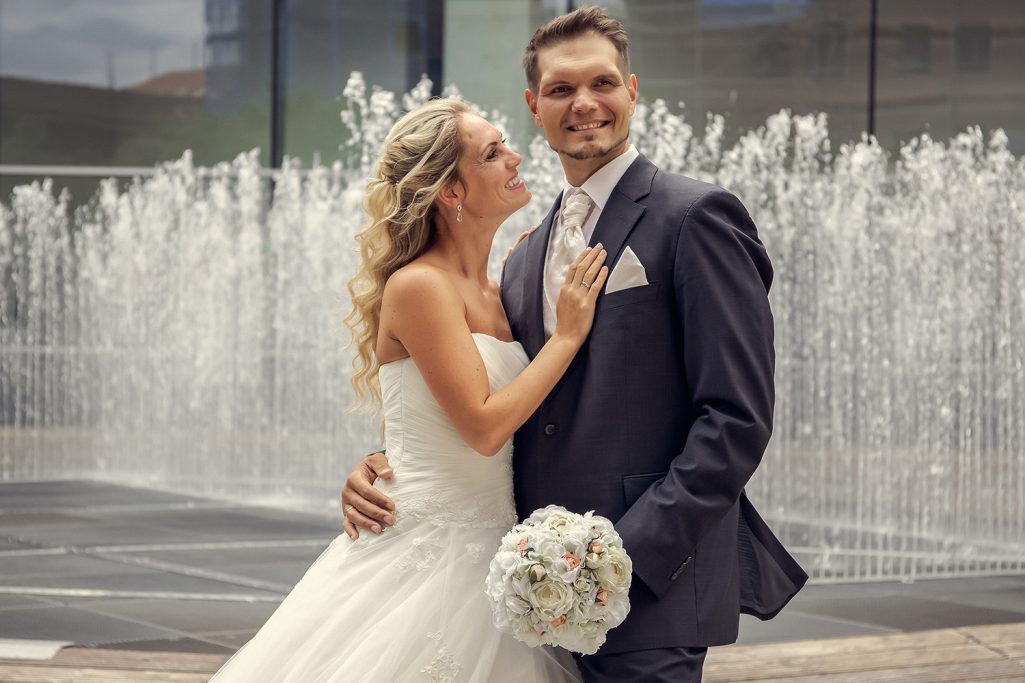 Ein Brautpaar posiert glücklich vor einem Wasserspiel. Die Braut lächelt den Bräutigam dabei an.