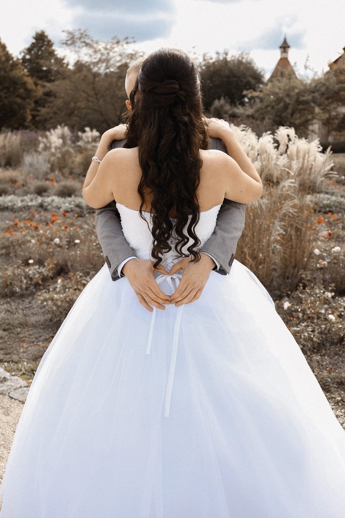 Hochzeitscouple aus der Rückansicht, Braut in weißem Kleid, der Bräutigam umarmt sie.