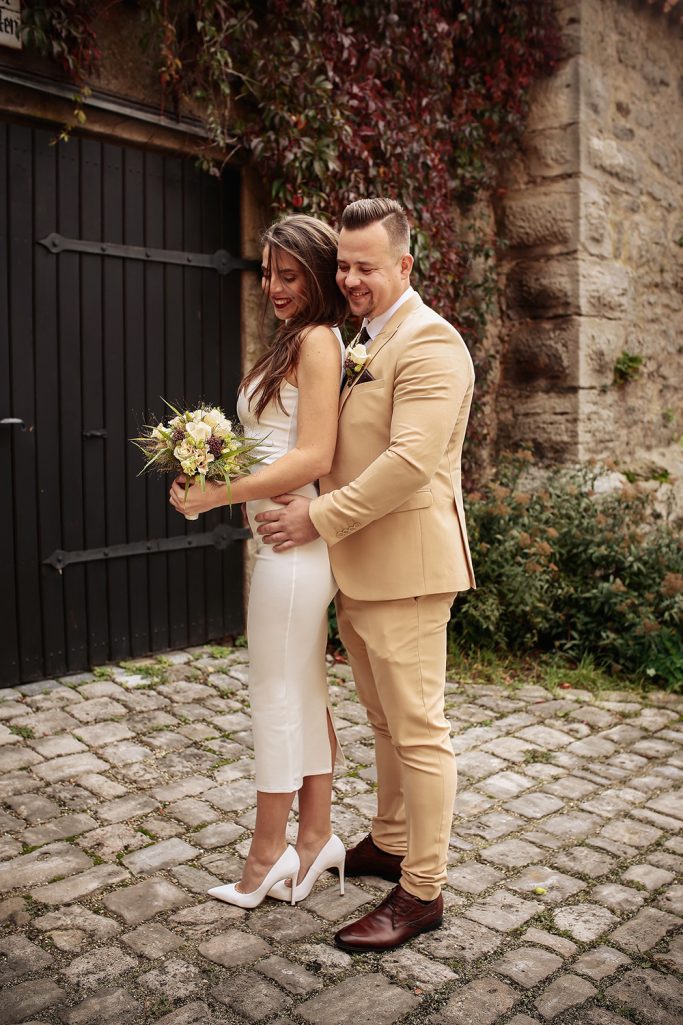 Brautpaar posiert vor einer steinernen Wand. Sie hält einen Blumenstrauß, er umarmt sie liebevoll.