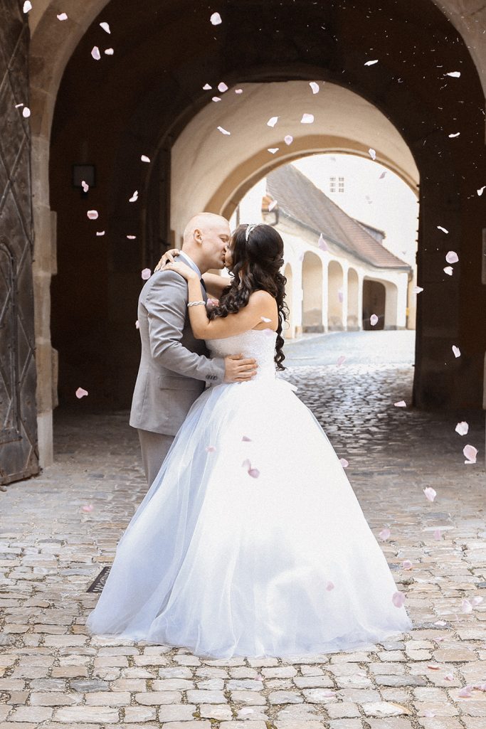 Braut und Bräutigam küssen sich unter einem Torbogen, umgeben von Blütenblättern.